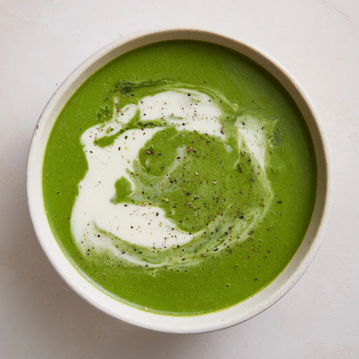 Steaming bowl of Crisp Spinach Soup with a swirl of cream and a sprinkle of black pepper beside a slice of crusty bread.