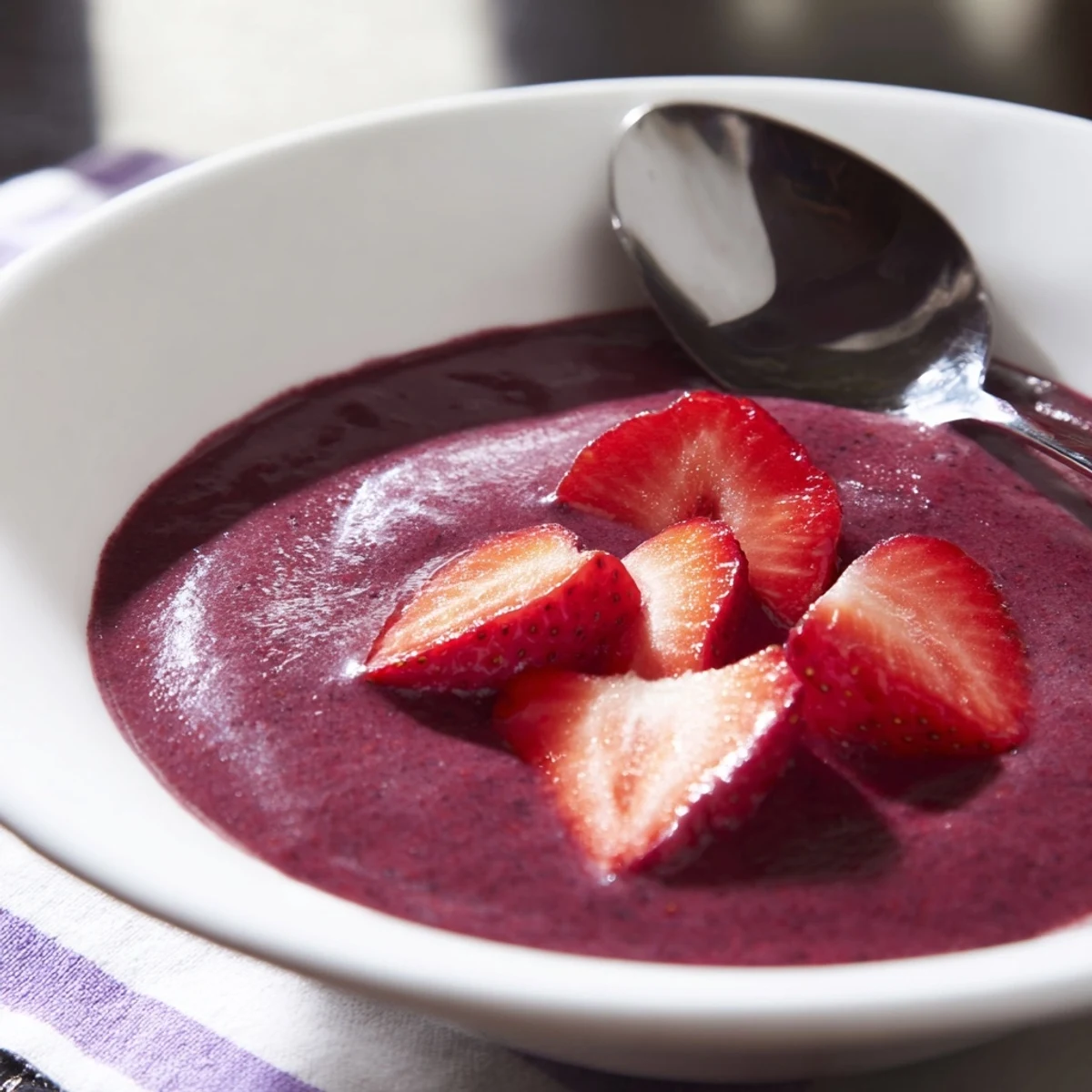 A chilled glass bowl displays vibrant Vegan Berry Soup, garnished with fresh strawberry slices, resting on a sunny windowsill for a refreshing summer treat.  