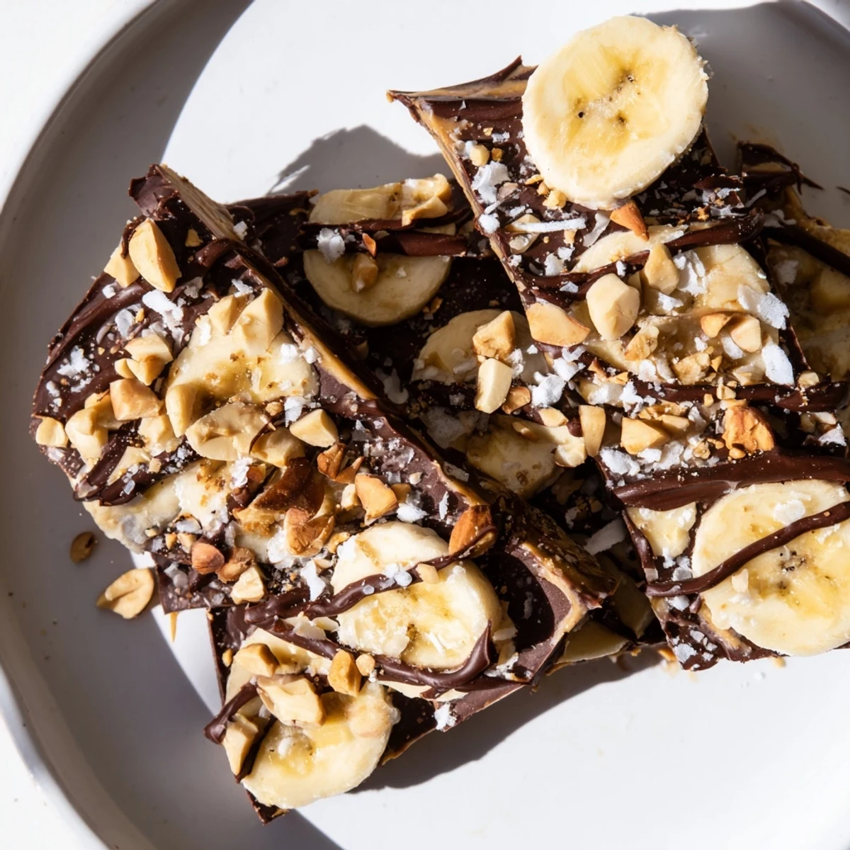 Frozen banana peanut butter bark drizzled with melted dark chocolate and flaky sea salt on white background